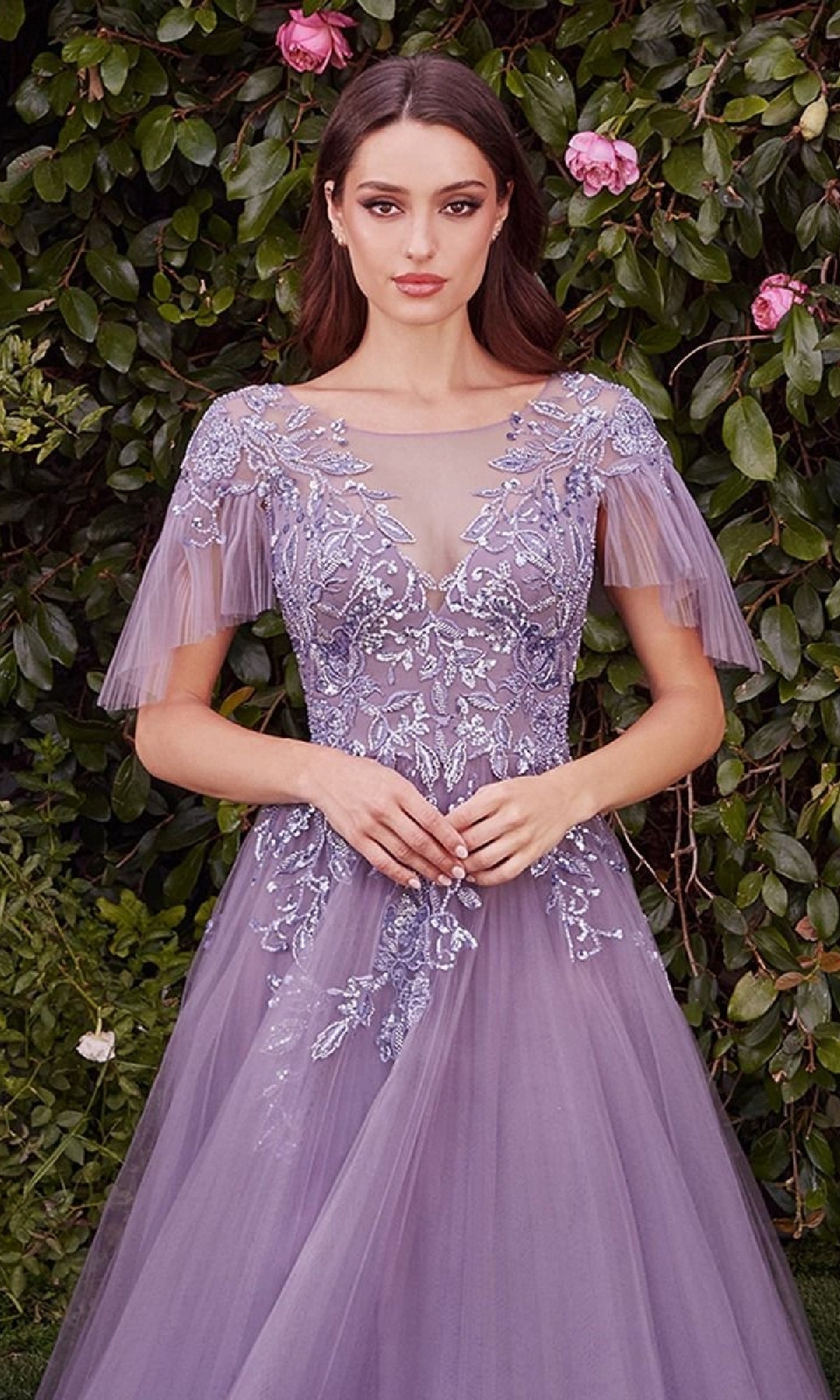 A woman stands in front of greenery wearing the Andrea & Leo A1351 lavender short-sleeve prom ball gown, featuring sheer sleeves and delicate floral embroidery.